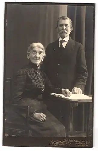 Fotografie E. Kregeloh, Mannheim, Breitestrasse, Portrait älteres Paar in hübscher Kleidung mit Buch am Tisch