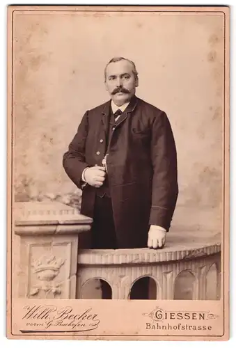 Fotografie Wilh. Becker, Giessen, Bahnhofstr., Portrait Herr im Anzug mit Walrossbart in einer Kulisse