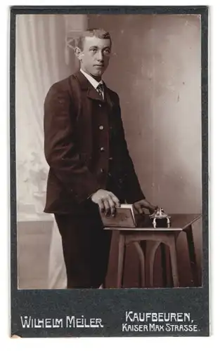 Fotografie Wilhelm Meiler, Kaufbeuren, Kaiser-Max-Strasse, Portrait junger Mann im Anzug mit Buch am Tisch stehend