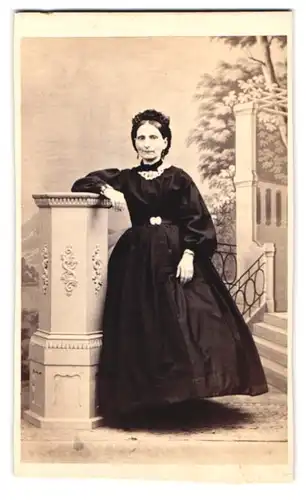 Fotografie unbekannter Fotograf und Ort, Portrait Dame im schwarzen reifrock Kleid mit Haube lehnt an einer Säule
