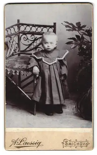 Fotografie A. Laessig, Smichov, Plzenska tr. 466, Portrait Kleinkind im dunklen Kleid steht an einem Stuhl gelehnt