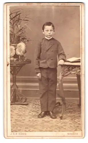 Fotografie J. C. H. König, Nossen, Bahnhofstrasse, Portrait frecher Bube im Anzug an einem Tisch stehend