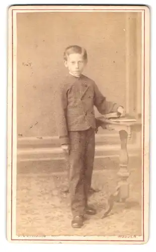 Fotografie J. C. H. König, Nossen, Bahnhofstr., Portrait frecher Bube im Anzug am Tisch stehend