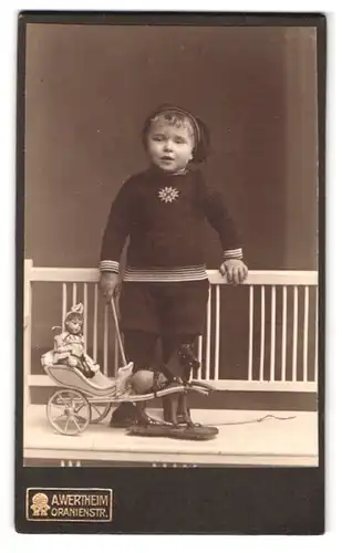 Fotografie A. Wertheim, Berlin, Oranienstr., Portrait junger Knabe im Anzug mit Zipfelmütze udn Puppenwagen
