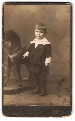 Fotografie Samson & Co., Essen / Ruhr, Portrait junger Knabe im Kleid mit Schaukelpferd und Spitzenkragen
