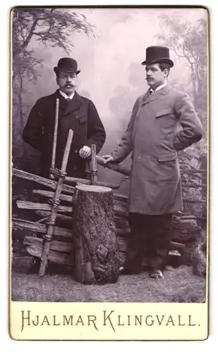 Fotografie Hjalmar Klingvall, Mora, Portrait zwei Herren in Anzügen und Melone, Zylinder, vor einer Studiokulisse