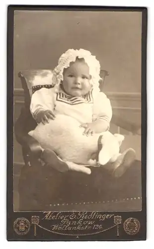 Fotografie E. Redlinger, Berlin-Pankow, Wollankstrasse 124, Portrait modisch gekleidetes Kleinkind mit Haube und Kissen