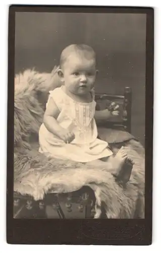 Fotografie D. Weissgärber, Zwönitz i. S., Bahnhofstrasse, Portrait niedliches Baby im weissen Kleidchen auf Fell sitzend