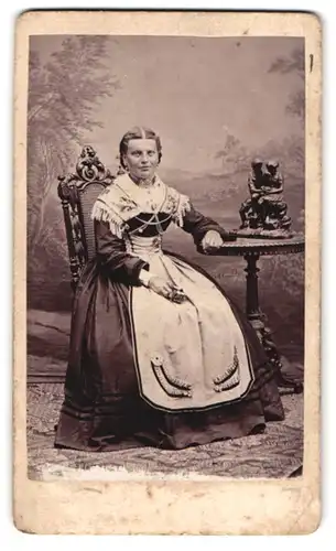 Fotografie J. Liesack, Rosenheim, Heiligengeiststr. 225, Portrait Dame im Trachtenkleid am Tisch mit Bronzestatuette