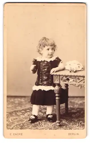 Fotografie E. Encke, Berlin, Potsdamerstr. 125, Portrait niedliches Mädchen im seidenen Kleid mit Puppe auf Tisch