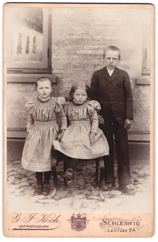 Fotografie G. J. Koch, Schleswig, Lollfuss 24, Portrait zwei kleine Mädchen in Kleidern mit einem Jungen