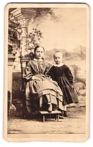Fotografie unbekannter Fotograf und Ort, Portrait zwei niedliche Mädchen in Kleidern vor einer Studiokulisse