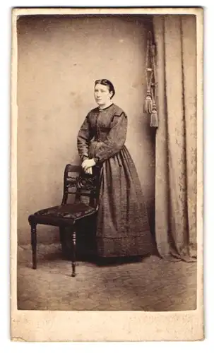 Fotografie unbekannter Fotograf und Ort, Portrait Dame im schlichten Biedermeierkleid mit Brosche steht hinterm Stuhl