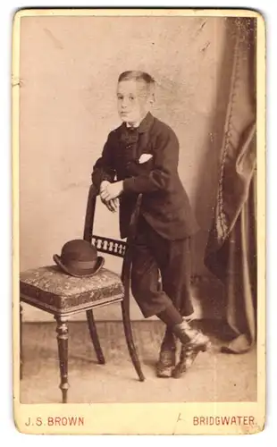 Fotografie J. S. Brown, Bridgwater, High Street, Portrait frecher Bube lehnt im Anzug lässig an einem Stuhl