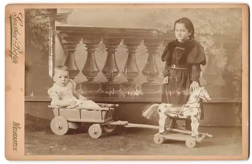 Fotografie Gustav Bopp, Weingarten, Portrait zwei Kinder mit Holzpferd und Wagen spielen im Atelier