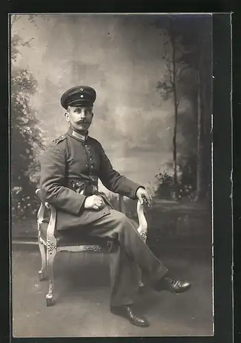 AK Soldat in Uniform mit Mütze auf einem Stuhl sitzend, Uniformfoto