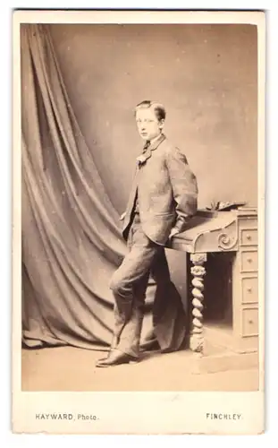 Fotografie Robert Hayward, Finchley, Portrait junger Knabe im grauen Anzug an einen Sekretär gelehnt