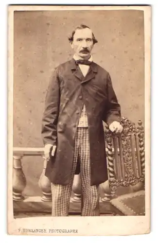 Fotografie Pierre Hunsanger, Ort unbekannt, Portrait Herr im karierten Anzug mit Mantel und Fliege, Moustache