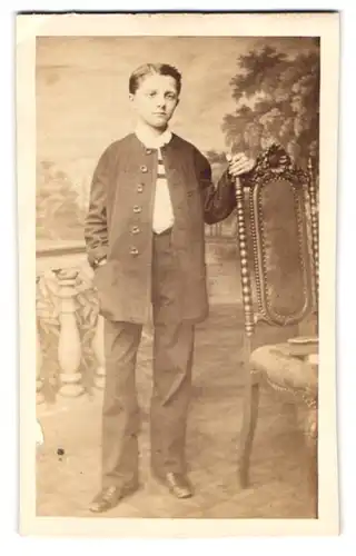 Fotografie unbekannter Fotograf und Ort, Portrait junger Knabe im Anzug steht neben einen Korkenzieherstuhl