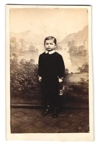 Fotografie unbekannter Fotograf und Ort, Portrait kleiner Knabe im schwarzen Anzug mit Sommerhut in der Hand