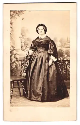 Fotografie unbekannter Fotograf und Ort, Portrait Dame im seidenen Biedermeierkleid mit Haube vor einer Studiokulisse