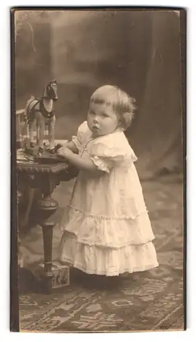 Fotografie unbekannter Fotograf und Ort, Portrait kleines Mädchen im weissen Kleid mit Holzpferd