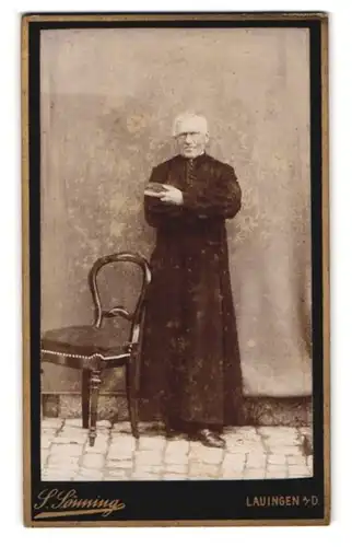 Fotografie S. Soenning, Lauingen, Portrait älterer Pastor im Talar mit Bibel in der Hand, lehnt an einer Wand