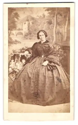 Fotografie unbekannter Fotograf und Ort, skeptisch blickende Dame im weiten Biedermeierkleid vor Studiokulisse