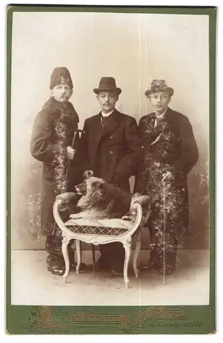 Fotografie Wilhelm Stein, Berlin, Portrait drei Herren in Wintermänteln mit Hund auf dem Stuhl