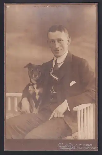 Fotografie Rud. Obigt, Berlin, Portrait junger Mann im Anzug mit seinem Hund im Arm auf einer Bank