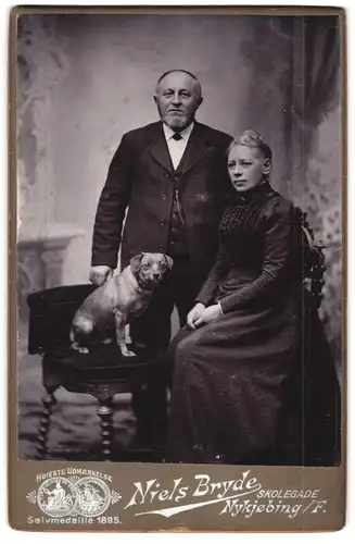 Fotografie Niels Bryde, Nykjöbing, Portrait älteres Paar im Biedermeierkleid und Anzug mit Hund im Studio