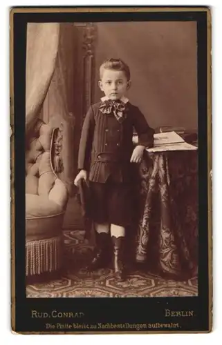 Fotografie Rud. Conrad, Berlin, Portrait junger Knabe im Anzug mit kurzen Hosen udn Schleife
