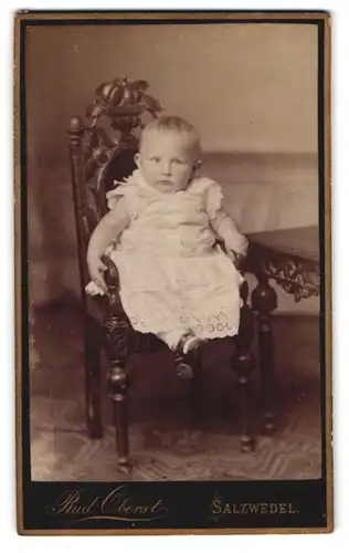 Fotografie Rud. Oberst, Salzwedel, Portrait Kleinkind im weissen Kleidchen auf einem Kinderstuhl