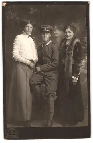 Fotografie Hermann Tietz, München, Bahnhofplatz, Soldat in Uniform mit Schirmmütze von Damen flankiert