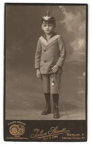 Fotografie Julius Staudt, Berlin, Unter den Linden 47, Knabe im Matrosenanzug mit Pickelhaube Preussen & Säbel
