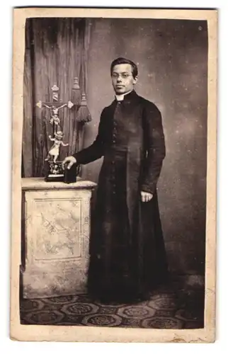 Fotografie unbekannter Fotograf und Ort, Portrait junger Pastor im Talar mit aufgestelltem Kreuz im Atelier