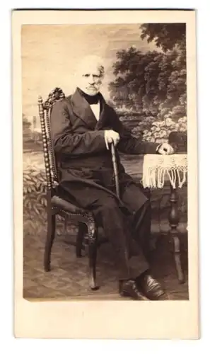 Fotografie Heinrich Fritz, Greiz, Portrait älterer Herr im Anzug mit Spazierstock sitzend im Atelier