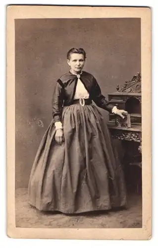 Fotografie Hermann Henning, Bornheim, Portrait Dame im Biedermeierkleid mit Bolero steht am Sekretär