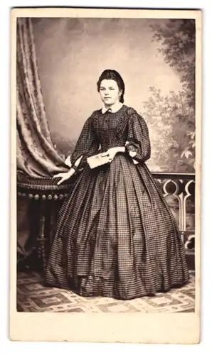 Fotografie unbekannter Fotograf und Ort, Portrait junge Frau im karierten Reifrock Kleid mit Fotoalbum in der Hand