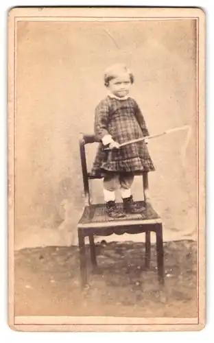 Fotografie unbekannter Fotograf und Ort, Portrait kleines Kind im karierten Kleid mit Peitsche