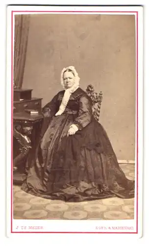 Fotografie J. Th. Meier, Eger, Portrait Frau Eger im weiten Biedermeierkleid mit Haube sitzend am Sekretär