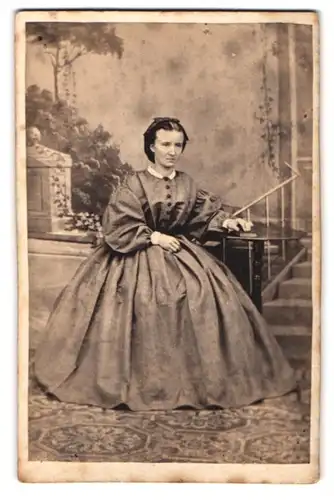 Fotografie unbekannter Fotograf und Ort, Hausfrau im Biedermeierkleid vor Studiokulisse sitzend