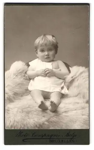 Fotografie Photo-Compagnie Belge, Bruxelles, 109, Rue Neuve, Portrait süsses Kleinkind im weissen Hemd sitzt auf Fell