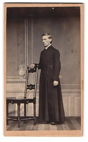 Fotografie unbekannter Fotograf und Ort, Portrait Pfarrer im Talar mit Collar stehend am Stuhl