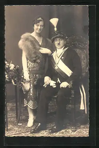 AK Mann mit extravaganter Kopfbedeckung, Schärpe und Orden geht mit seiner Frau zum Fasching