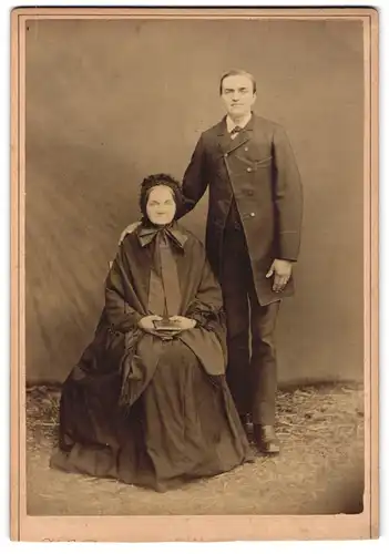 Fotografie unbekannter Fotograf und Ort, Portrait ältere Dame im Umhang mit Haube und junger Herr