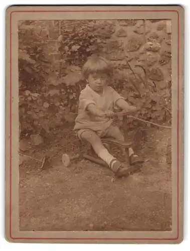 Fotografie unbekannter Fotograf und Ort, Portrait junger Knabe auf seinem Dreirad im Garten