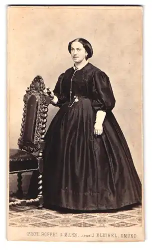 Fotografie Boppel & Mahn, Schwäbisch Gmünd, Portrait Dame im dunklen reifrock Kleid stehend am Stuhl