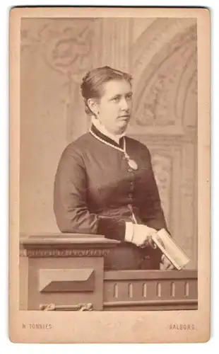 Fotografie H. Tönnies, Aalborg, Portrait junge Dame im Kleid mit Amulett und Buch