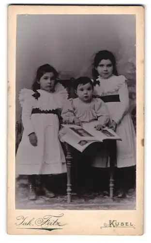 Fotografie Jakob Fritz, Kusel /Pfalz, Bahnhofstrasse, Portrait süsses Kleinkind im Kleid mit Buch und zwei Mädchen